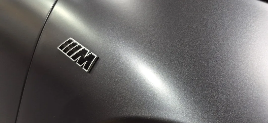 BMW iX M70 badge with matte paintwork in background